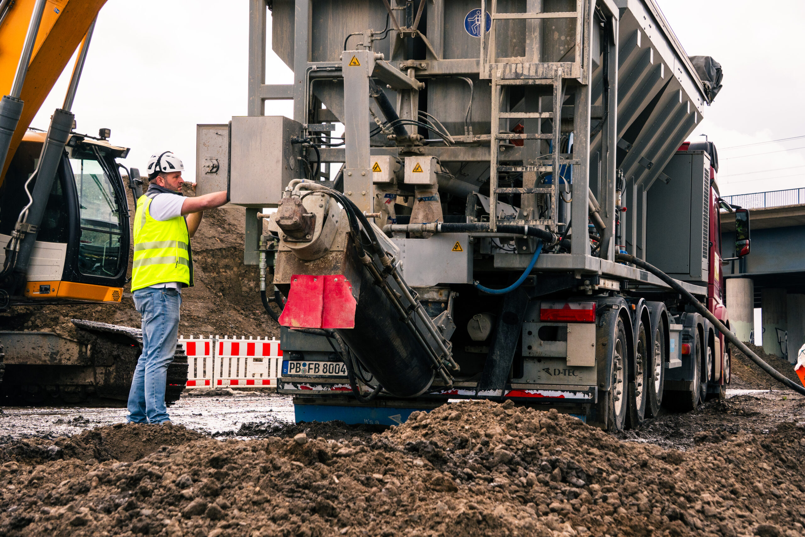 Mobile Flüssigbodenmischanlage produziert Flüssigboden direkt auf einer Tiefbaustelle mit Bagger und Baustellenteam.